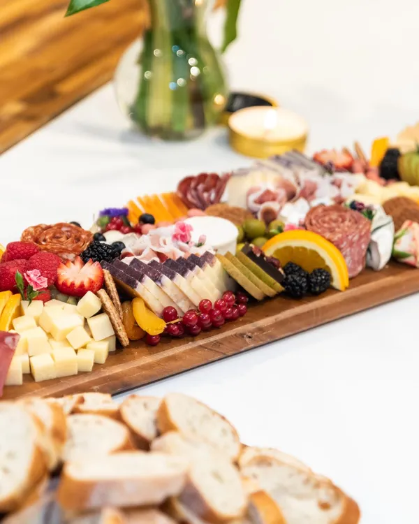 A long wooden board of cured meats, cheeses, fruit and edible flowers beside sliced baguette